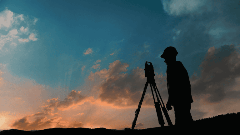 Surveying professional using a theodolite during sunset for land measurement.