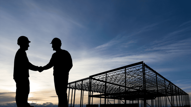 Two workers shaking hands at a construction site with steel framework in the background.